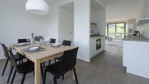 Une cuisine moderne avec des meubles blancs et un îlot central, un coin repas avec une table en bois et des chaises noires, et une vue sur un balcon doté d’une balustrade.