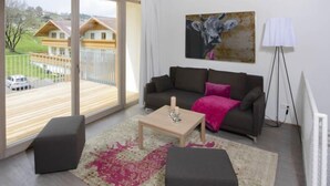 Un salon moderne avec un canapé, une table basse et un pouf, une grande fenêtre donnant sur des maisons et un tableau représentant une vache.