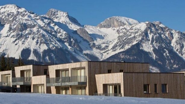 Ein modernes Gebäude mit großen Fenstern und einem Balkon, vor einer Kulisse schneebedeckter Berge.