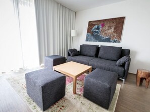Uma sala de estar moderna com um sofá cinzento, duas poltronas combinando, uma mesa de centro de madeira e um banquinho de madeira. Um quadro está pendurado na parede, e uma luminária está sobre uma mesinha lateral.