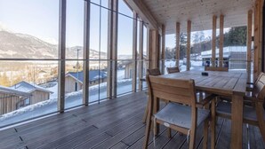A wooden deck with a table and chairs, large windows offering a view of a snowy landscape and buildings. - Superior 3 SZ Sauna & Aussenbadewanne by Interhome (Haus)