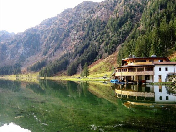 A lakeside cabin with a wooden deck and a boat dock, surrounded by mountains and dense forest.