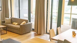 A modern living room with a sofa, a dining table, and chairs. There are large windows with curtains, and a view of the outdoors.