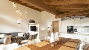 Ein modernes Wohnzimmer mit Holzboden, einem Essbereich mit großem Tisch, weißen Stühlen und einer Küche im Hintergrund.