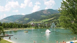 Ein Badestrand am See mit badenden und sonnenden Personen, umgeben von grünen Hügeln und einer Kirche.