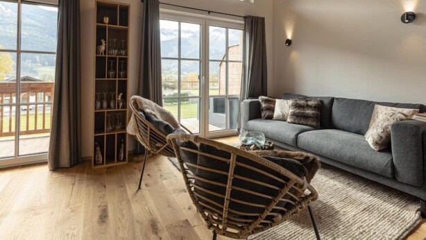 Ein modernes Wohnzimmer mit einem grauen Sofa, Korbstühlen, Holzboden und großen Fenstern mit Blick ins Freie.
