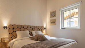 Een slaapkamer met een hoofdbord met houtpatroon, een groot bed met meerdere kussens, een nachtkastje met een lamp en een raam met uitzicht op gebouwen.