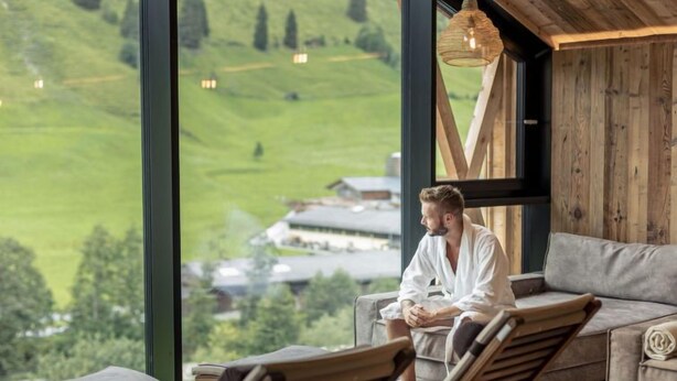 Eine Person in einem weißen Bademantel sitzt auf einer Liege vor einem großen Fenster mit Blick auf eine grüne Landschaft.