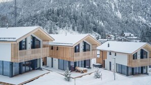 Een moderne chalet met een houten gevel en grote glasramen, omgeven door besneeuwde bergen.