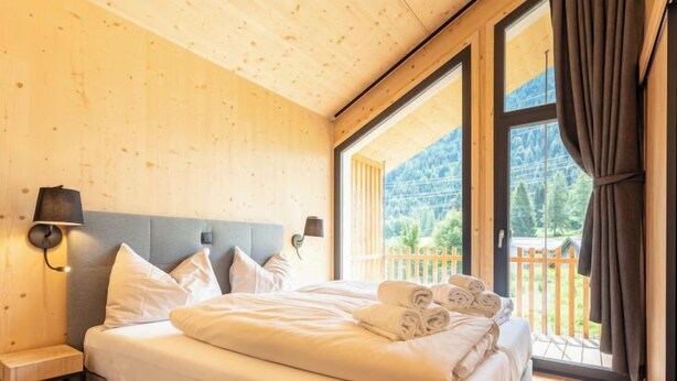Een slaapkamer met een groot bed, houten wanden, uitzicht op een houten terras en bergen, en een raam met gordijnen.