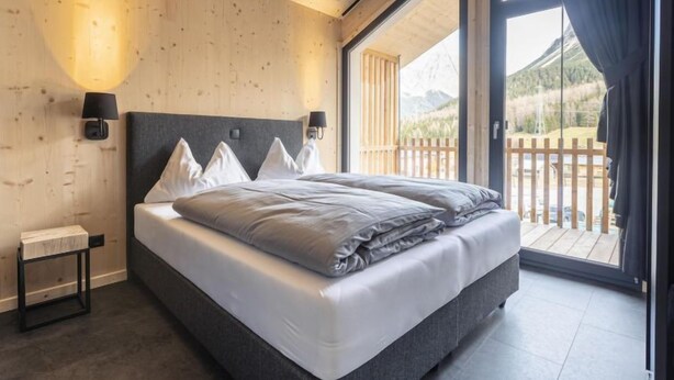 Een moderne slaapkamer met een groot bed, houten wanden en uitzicht op een balkon met houten hek en bergen op de achtergrond.