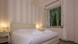 Uma cama bem arrumada com roupa de cama branca e um edredom listrado, uma mesa de cabeceira com abajur e uma janela com cortinas.