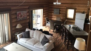 Interior acogedor de una cabaña de madera con sofá blanco, mesa de comedor, sillas, zona de cocina y un ventanal grande.