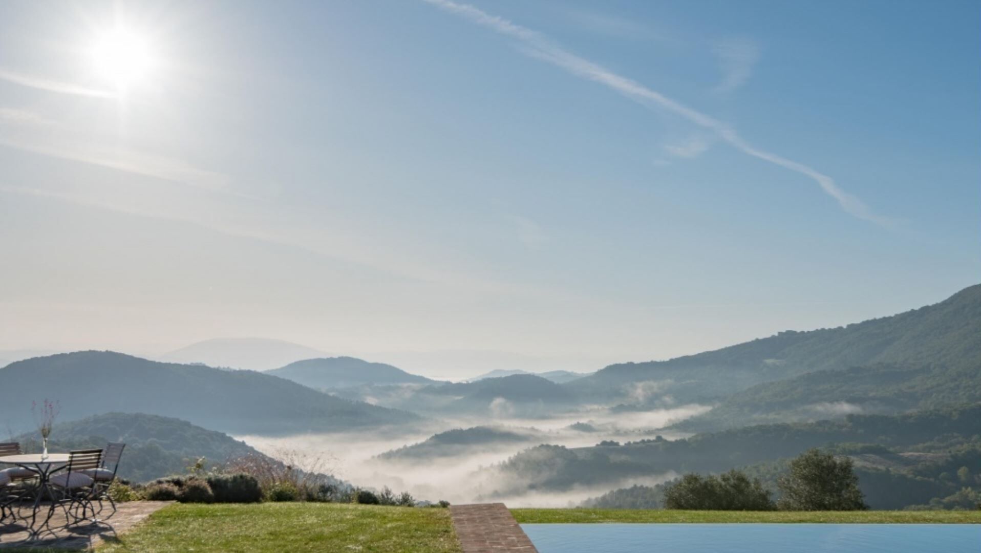 Ein malerischer Blick auf eine Berglandschaft mit blauem Himmel, einem Gewässer und einem Tisch mit Stühlen auf einer Wiese.