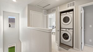 A laundry room with two stacked front-loading washing machines, a door leading to a bedroom, and a small window with a view of a lawn. - Trendy Townhome 20 minutes to Fort Collins, Greeley, and Windsor (Ault)