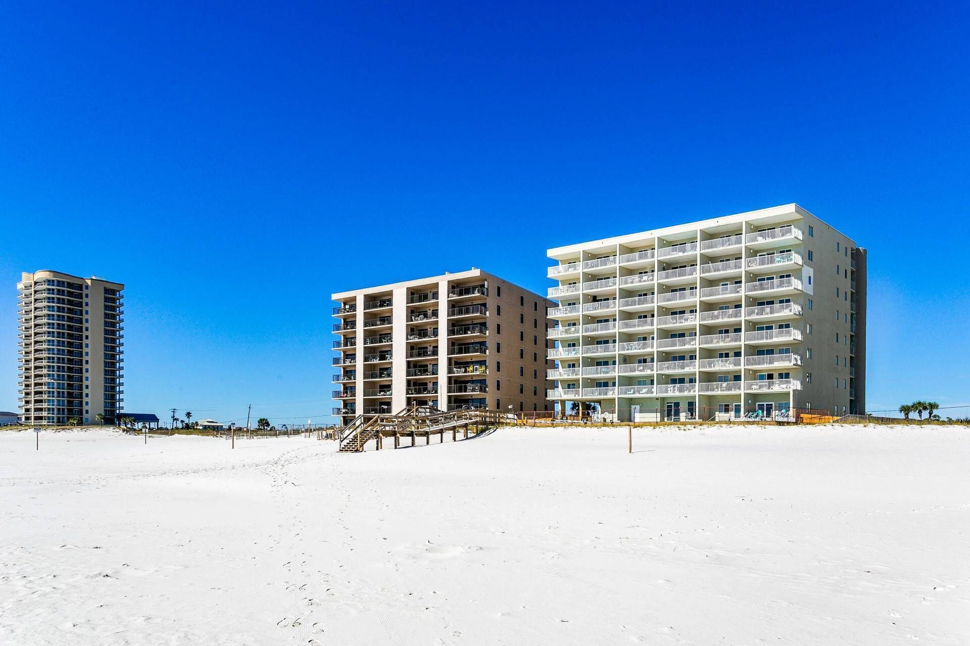 A beachfront hotel with multiple floors, a staircase leading to the beach, and a clear blue sky.