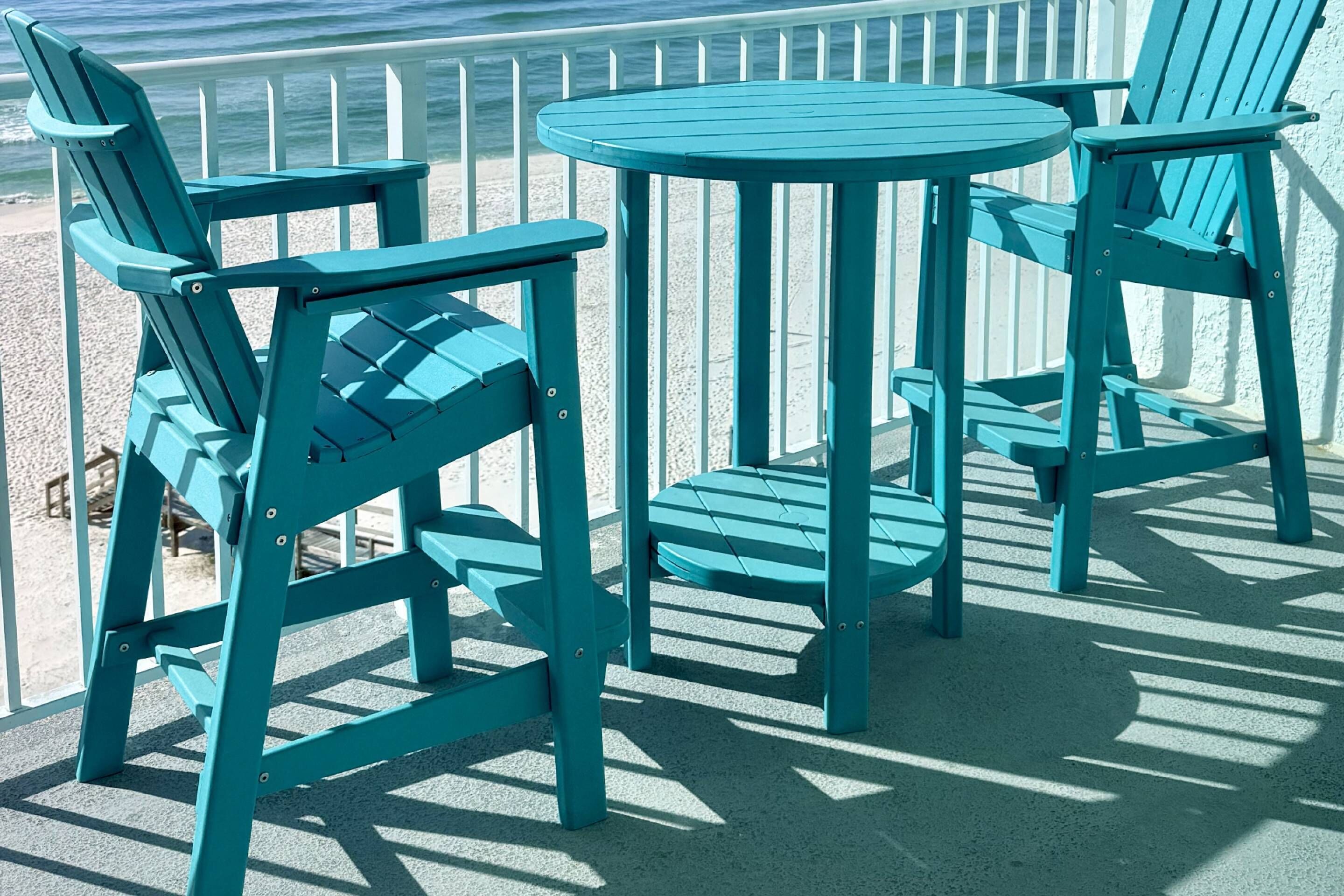 A set of teal Adirondack chairs and a round table on a balcony with a view of the ocean.