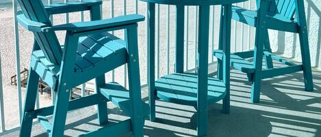 A set of teal Adirondack chairs and a round table on a balcony with a view of the ocean.
