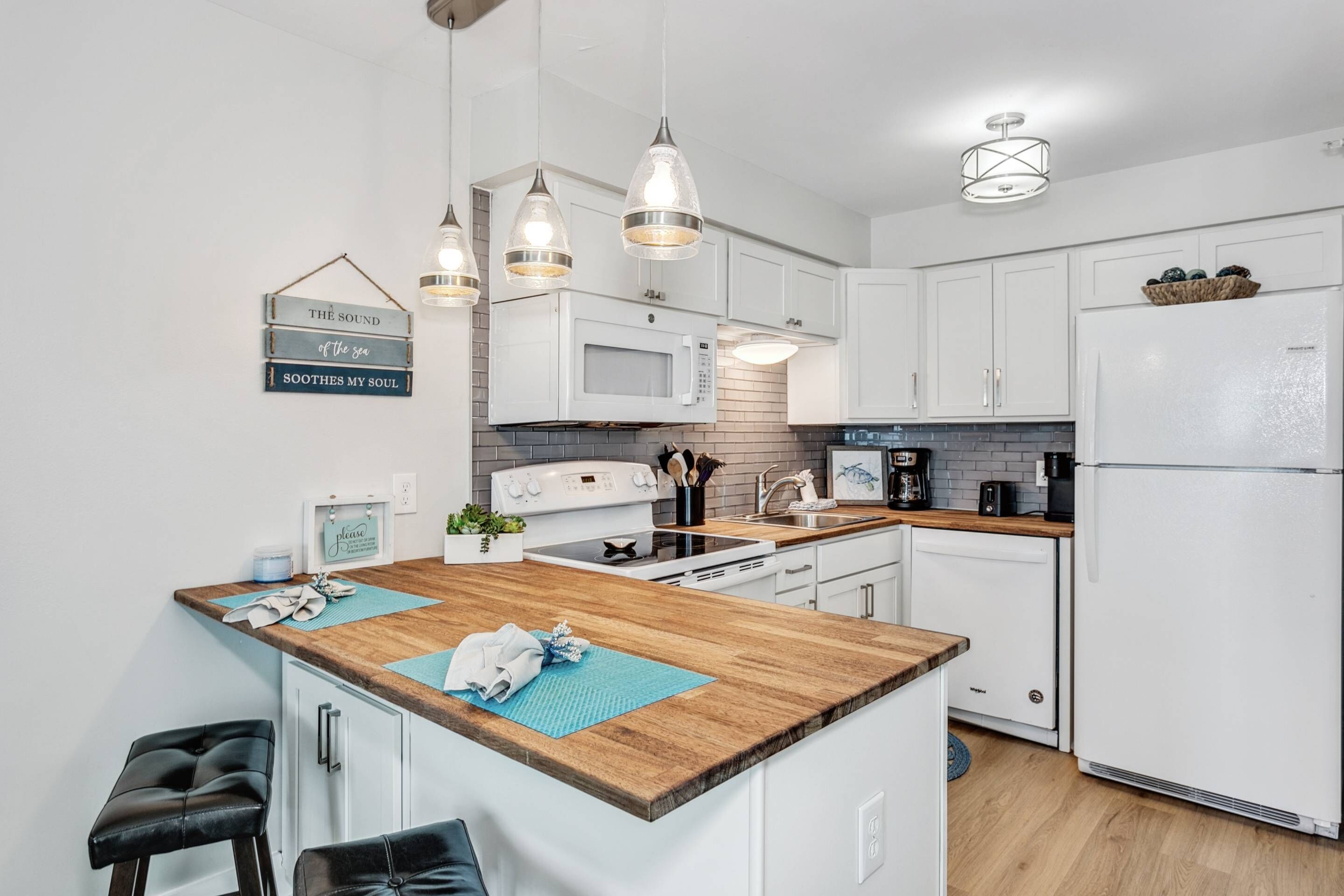 Une cuisine moderne avec des meubles blancs, un îlot central en bois, des luminaires suspendus et un réfrigérateur.