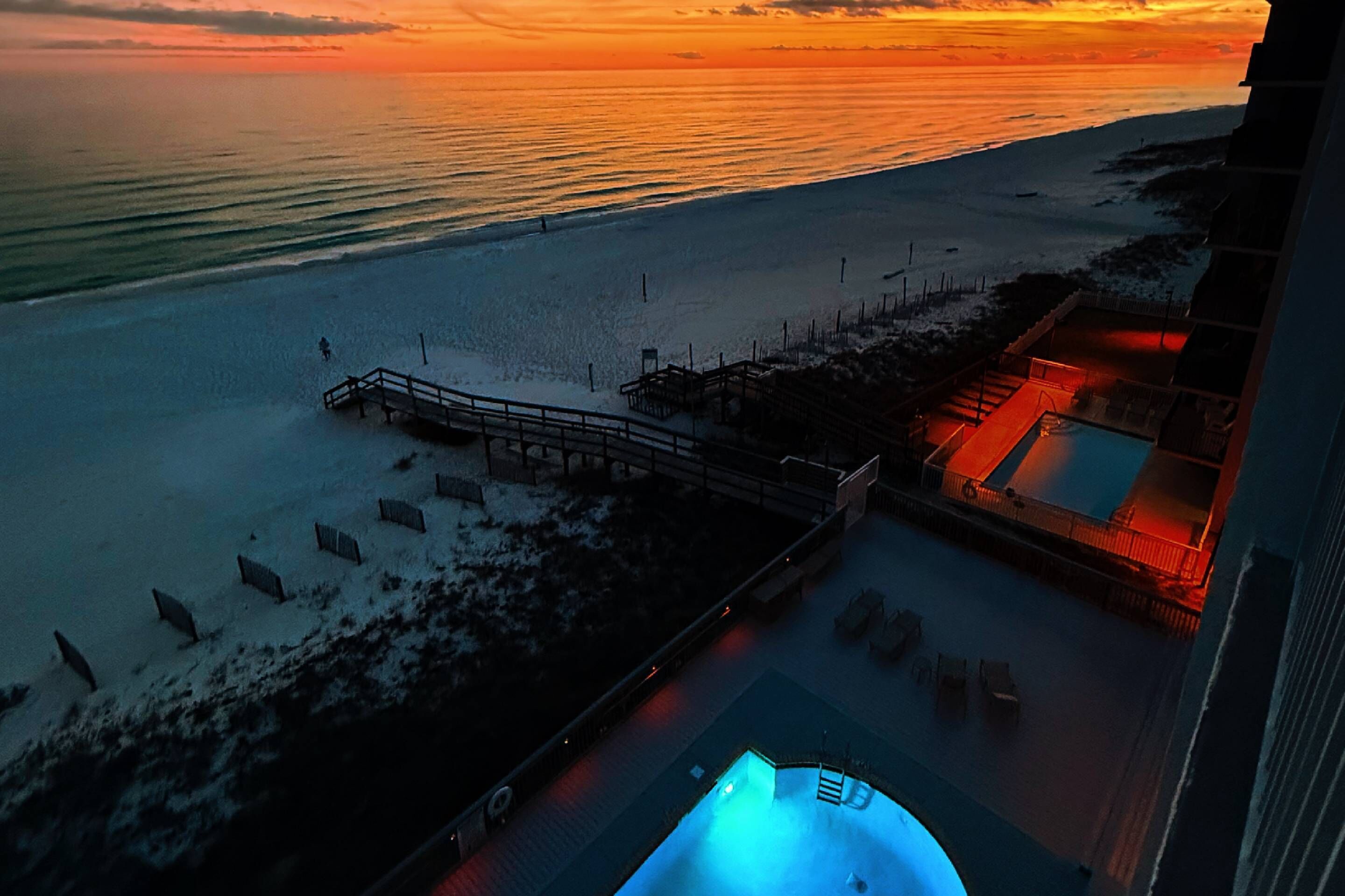 Vue aérienne d’un complexe hôtelier en bord de mer, avec une piscine, une terrasse en bois et une vue sur l’océan au coucher du soleil.