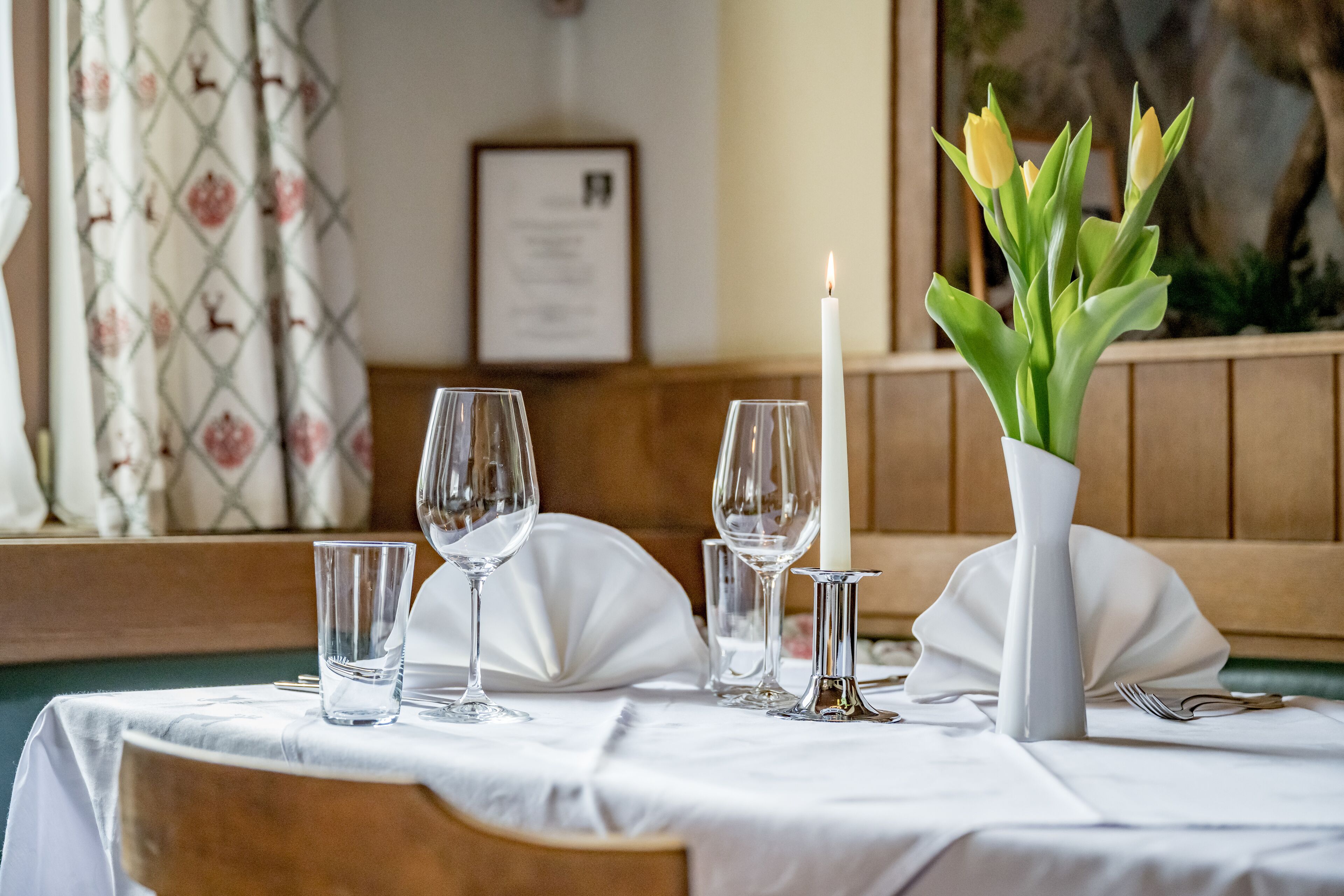 A dining table set with white tablecloth, clear glasses, a white candle, and a vase with green leaves.