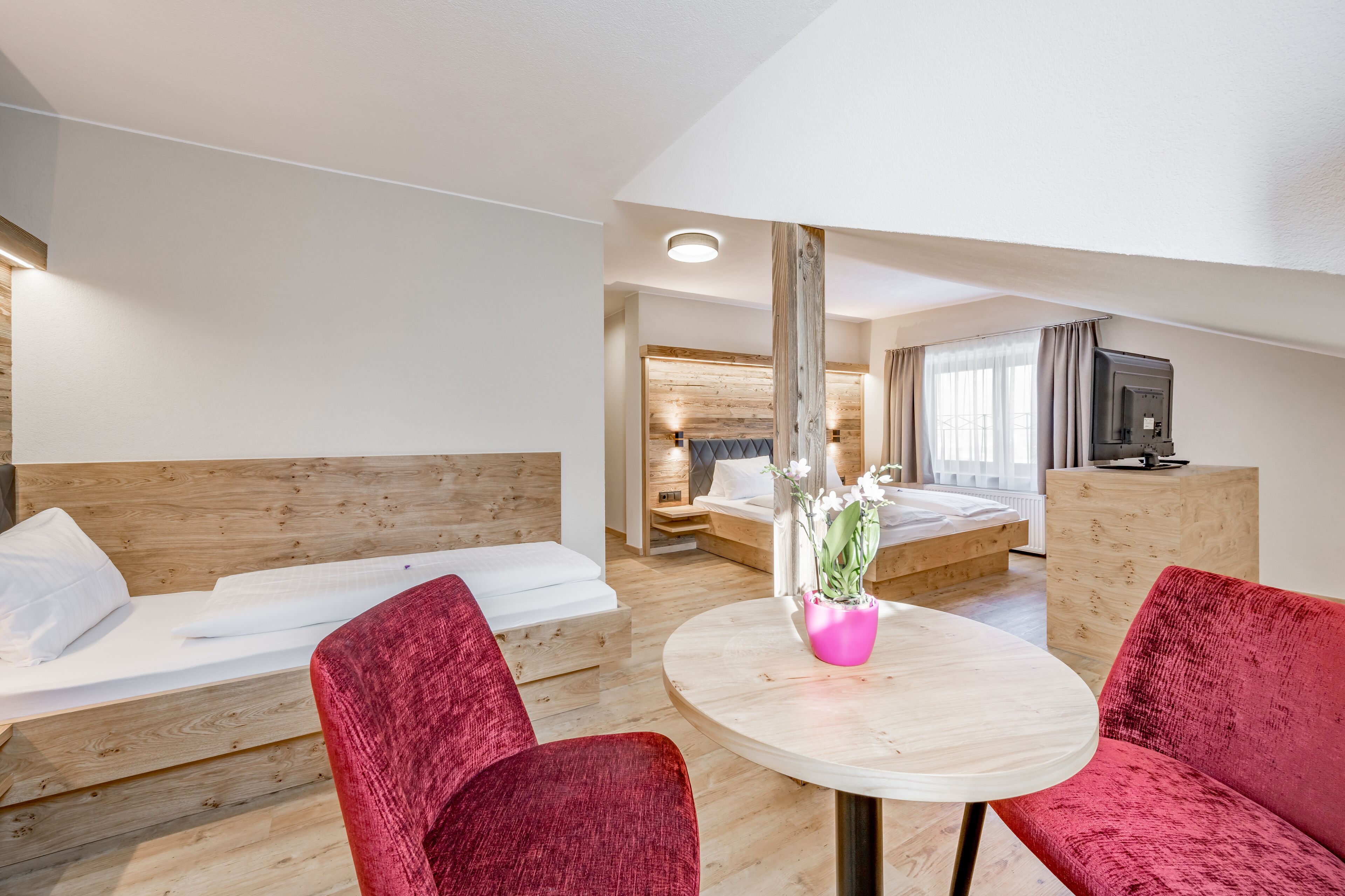 A modern hotel room with a wooden headboard, a round table with a pink vase, red upholstered chairs, a bed with white bedding, and a television on a wooden cabinet.