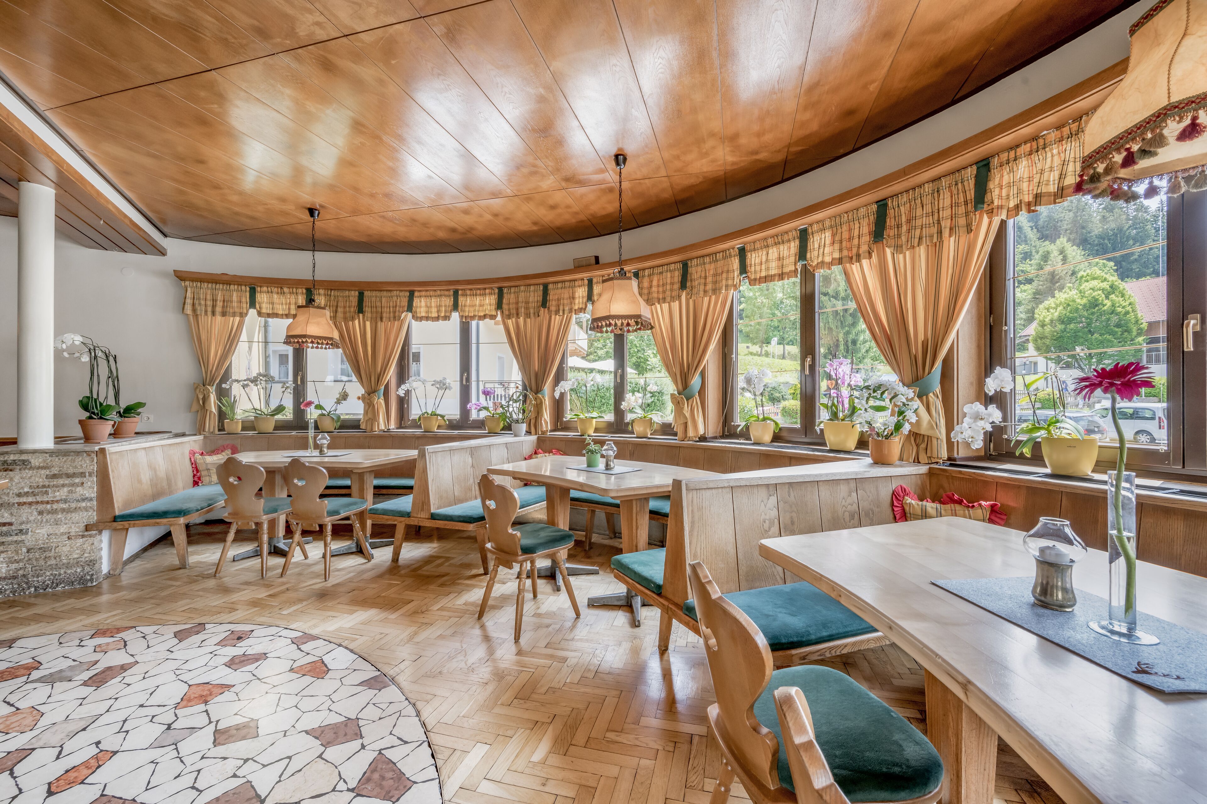 Ein gemütliches Restaurant mit Holztischen, großen Fenstern mit Vorhängen und Zimmerpflanzen.
