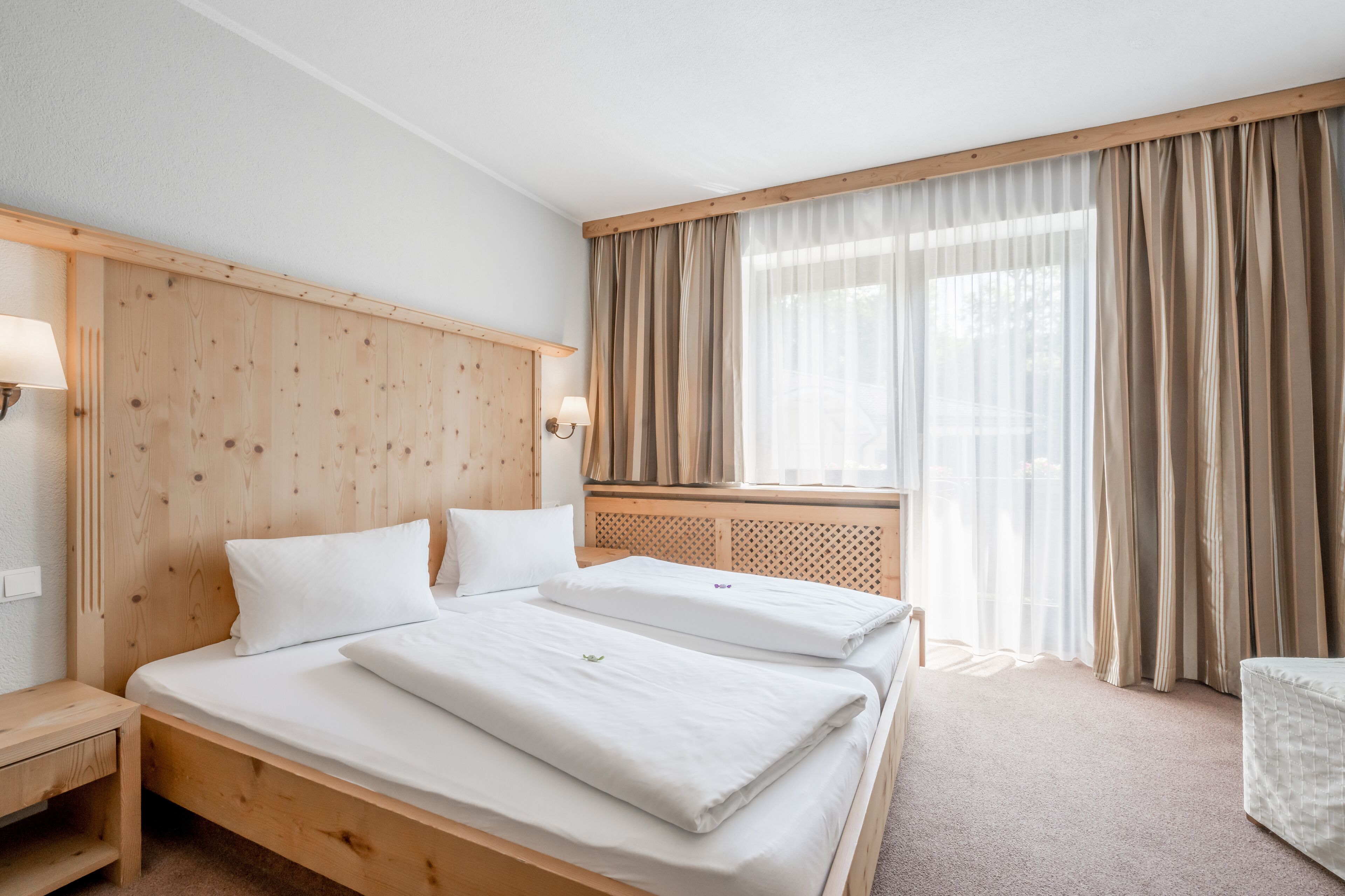 A hotel room with a wooden bed, two pillows, a nightstand, a lamp, and a window with curtains.