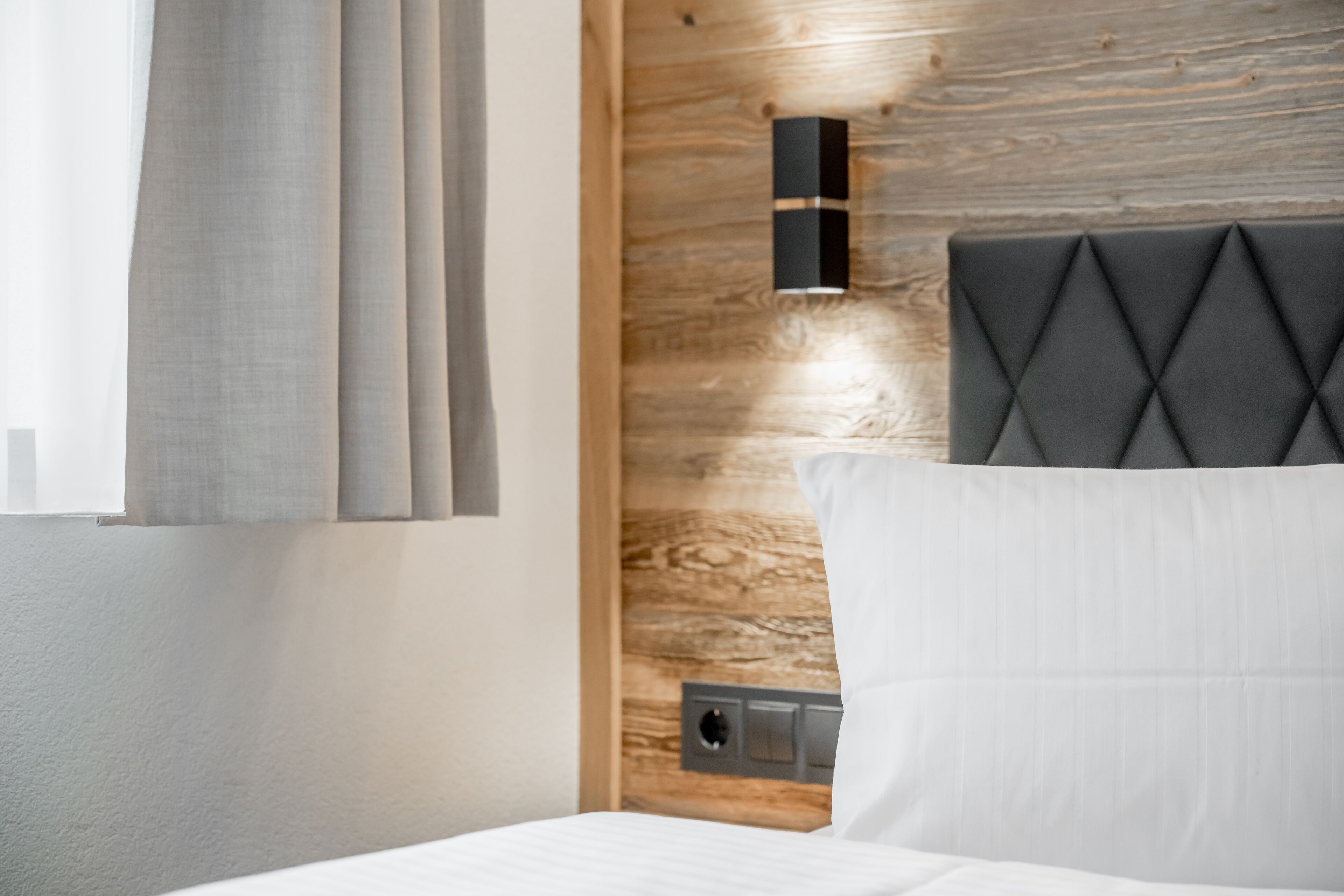 Une chambre d’hôtel moderne avec une tête de lit en bois, une table de chevet et une fenêtre avec des rideaux.