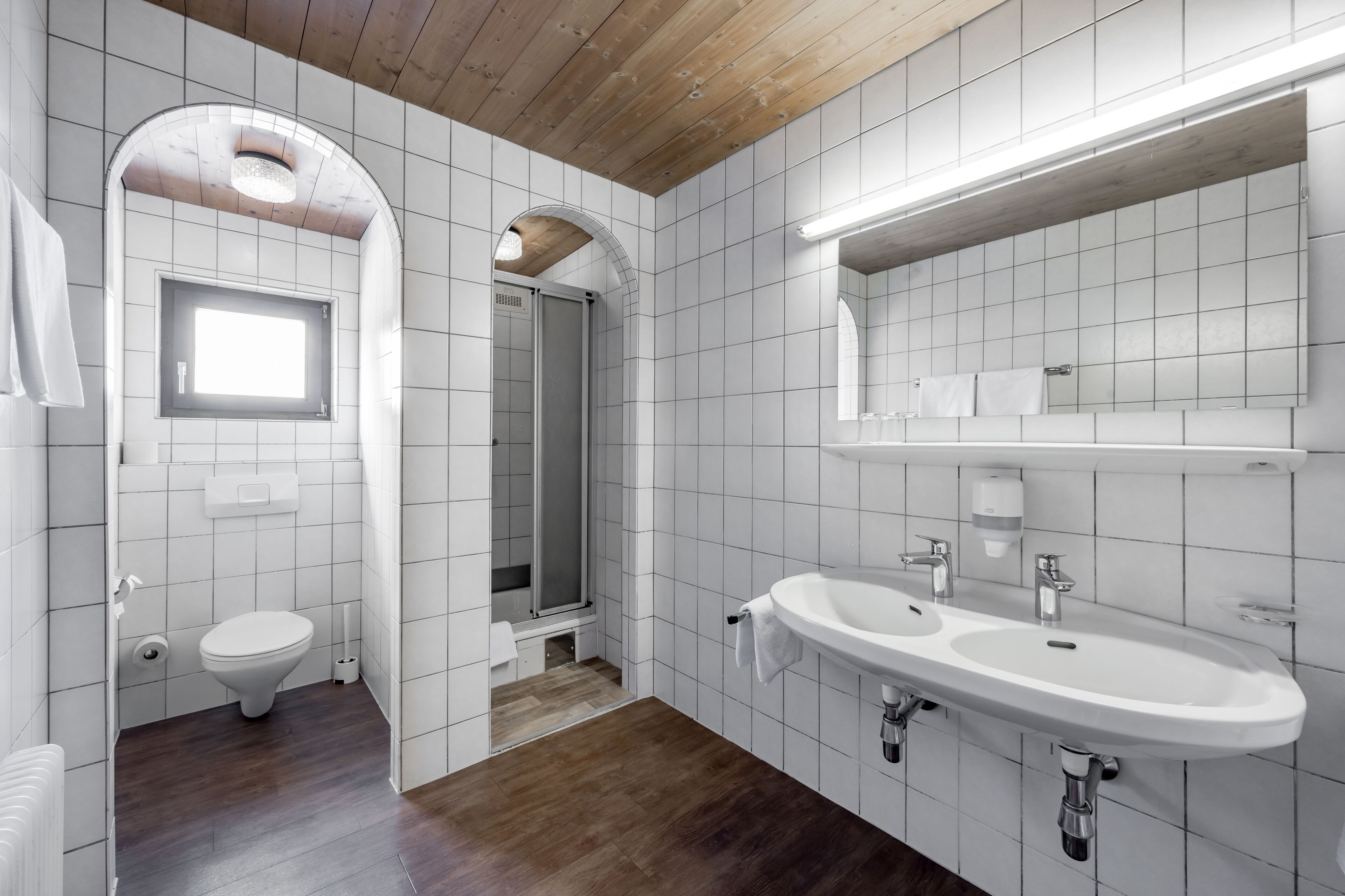 A modern bathroom with white tiles, a wooden ceiling, a large sink, and a shower area.