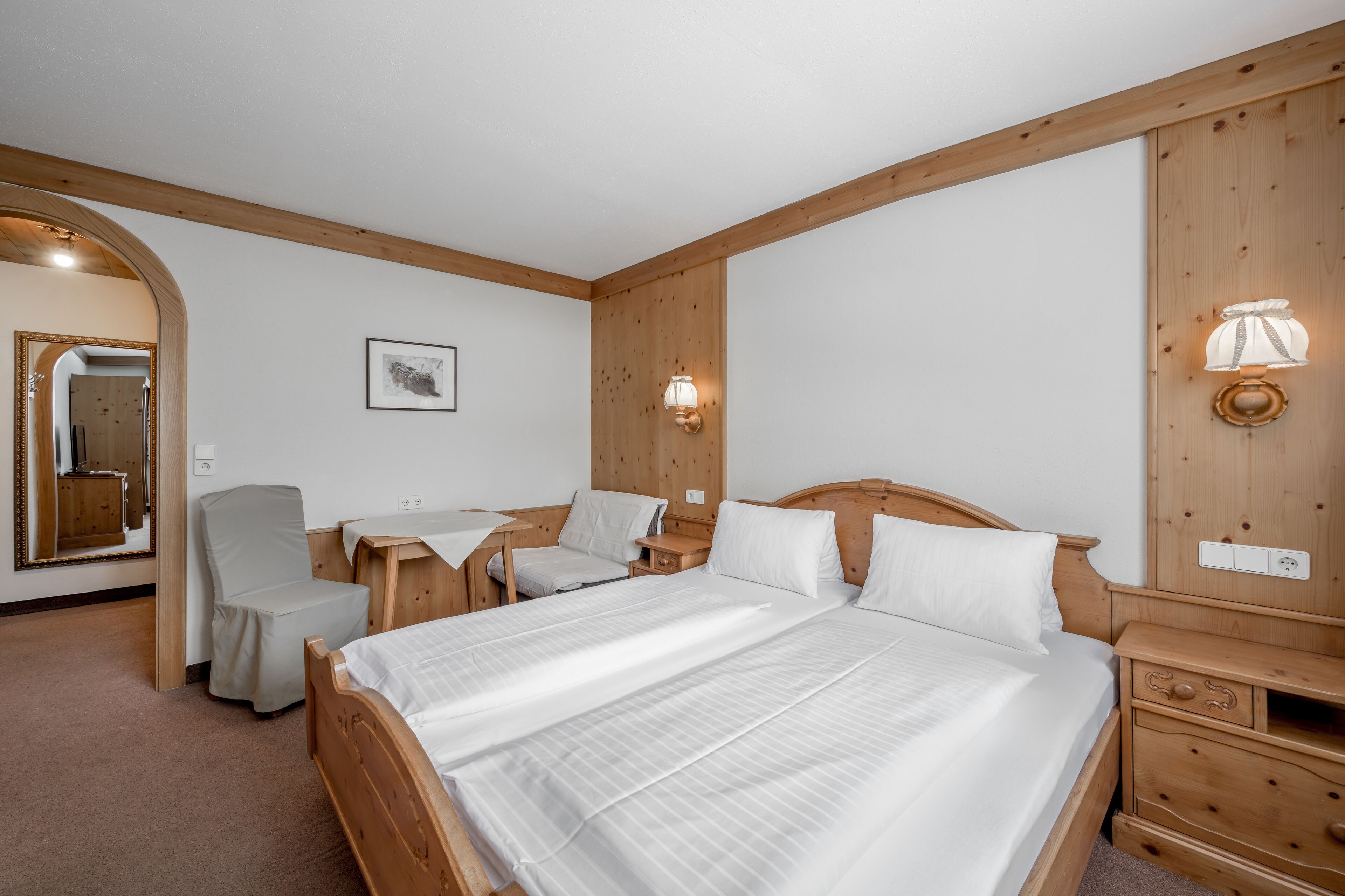 A hotel room with a large bed, a wooden wardrobe, a desk with a chair, and a mirror.