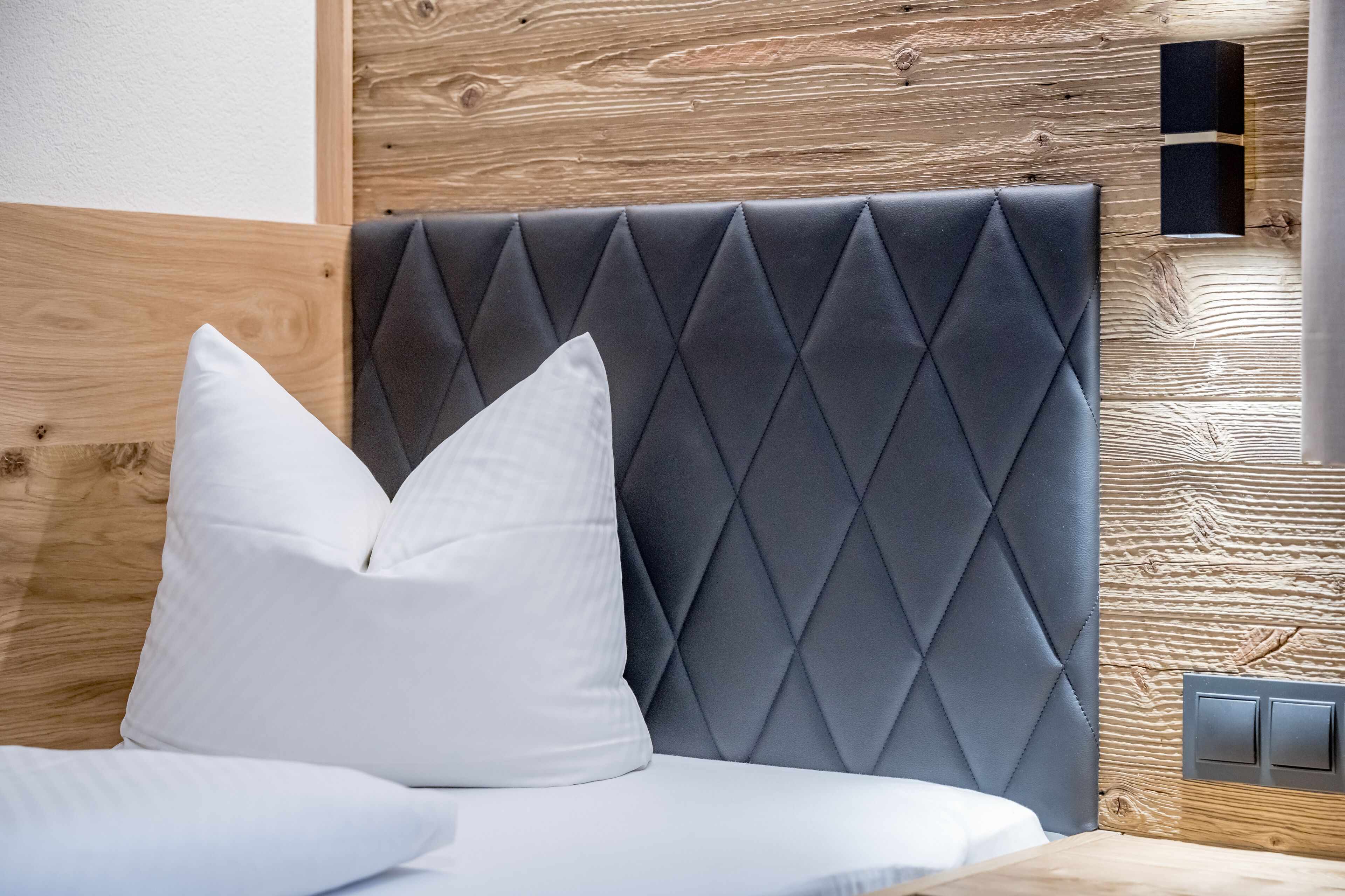 Ein Bett mit dunklem Kopfteil, zwei weißen Kissen, einer Holzpaneele und einem Lichtschalter.