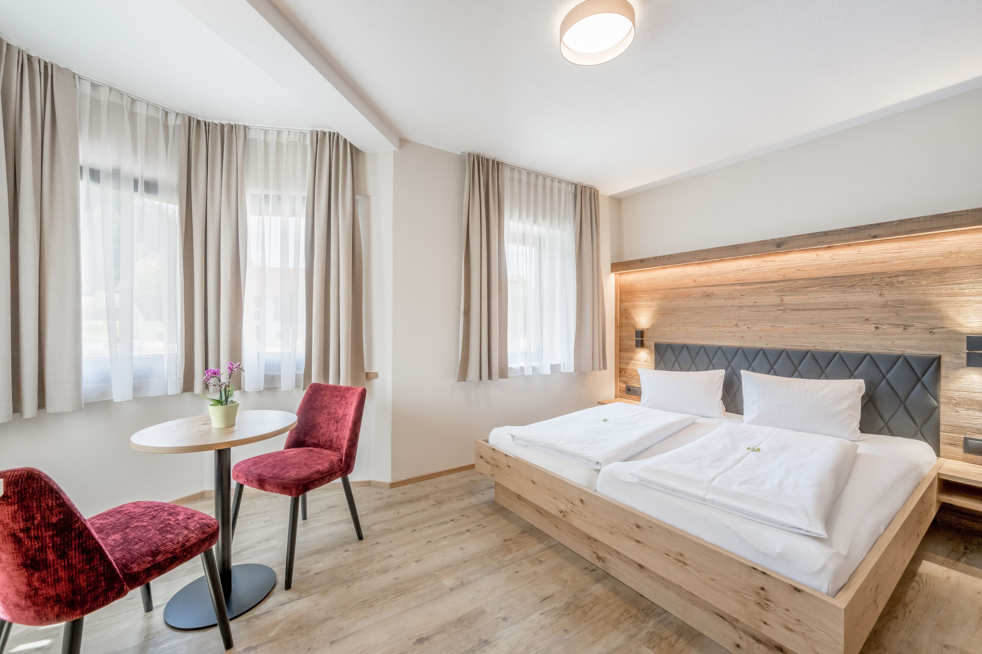 A hotel room with a wooden bed, two red chairs, a small round table, and large windows with curtains.