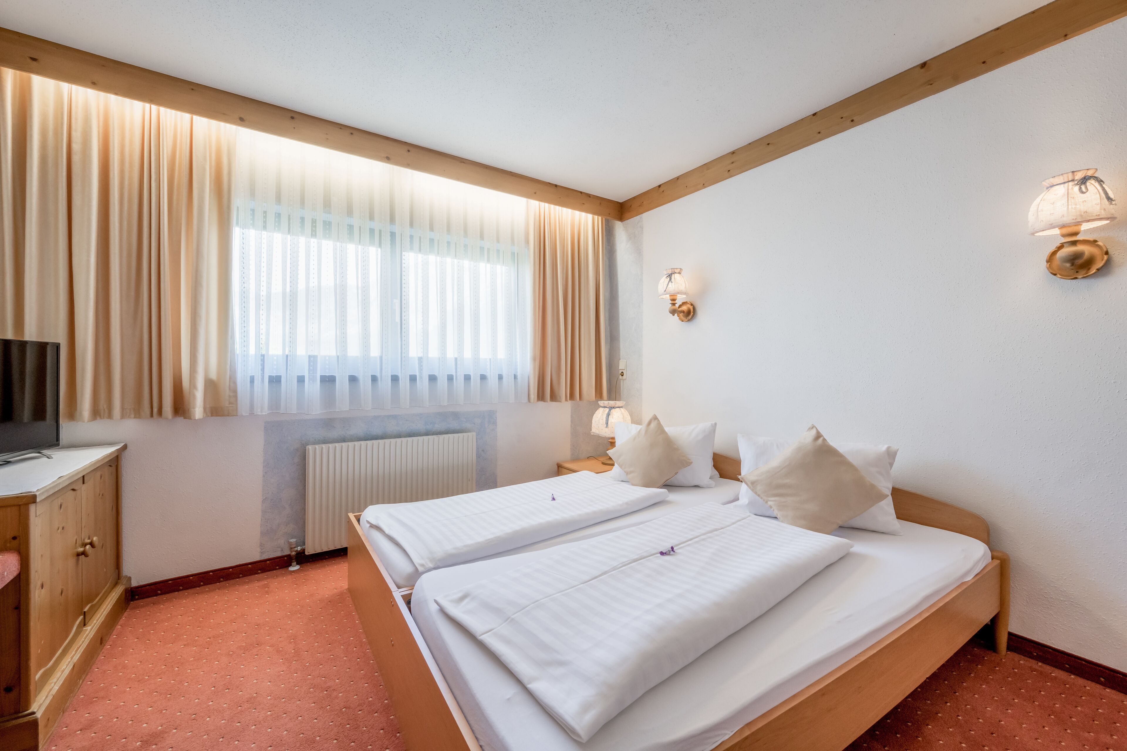 A hotel room with two beds, a wooden headboard, a TV, a radiator, and a window with curtains.