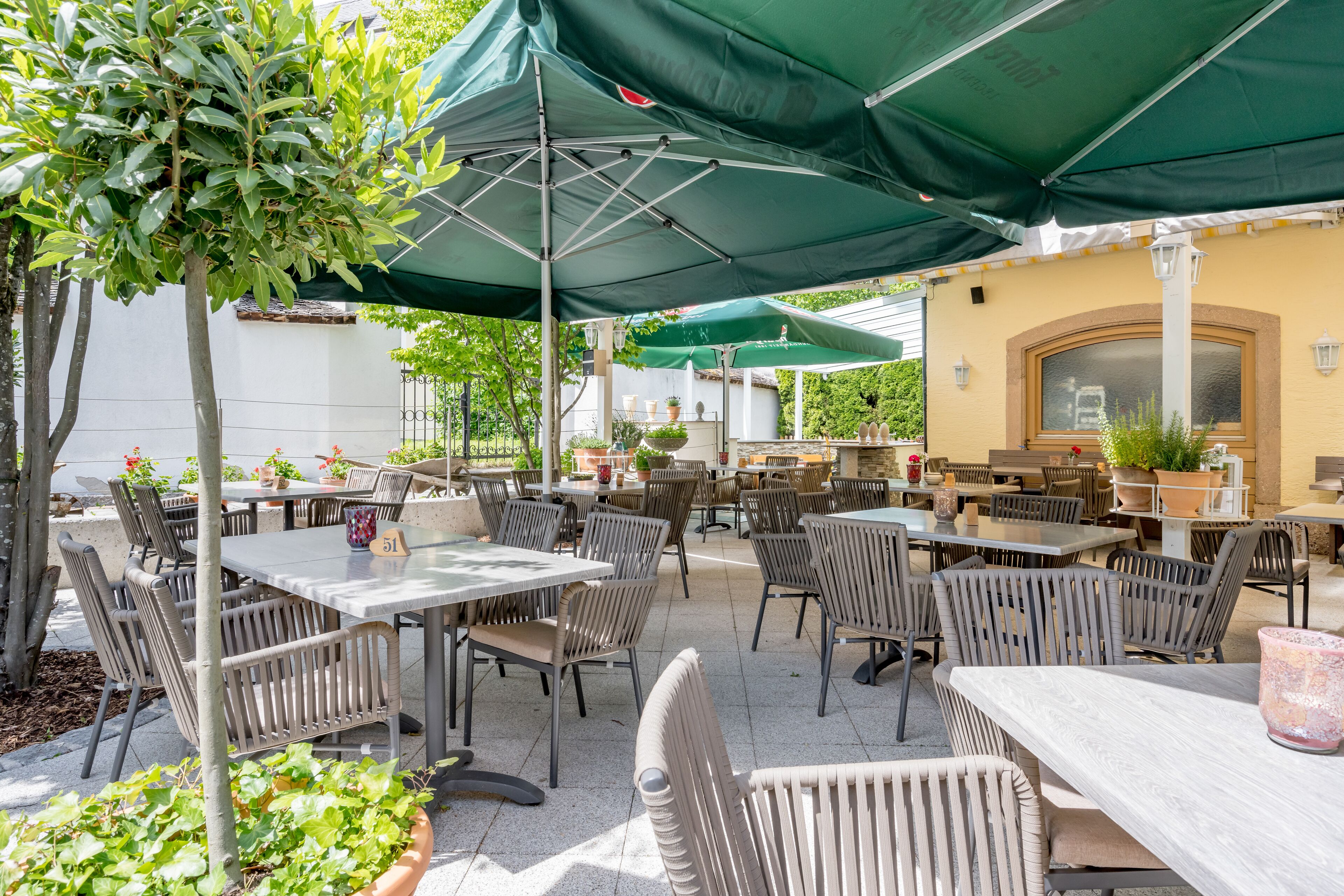 Ein Außenbereich mit Terrasse, Tischen, Stühlen, grünen Sonnenschirmen und Blumentöpfen.