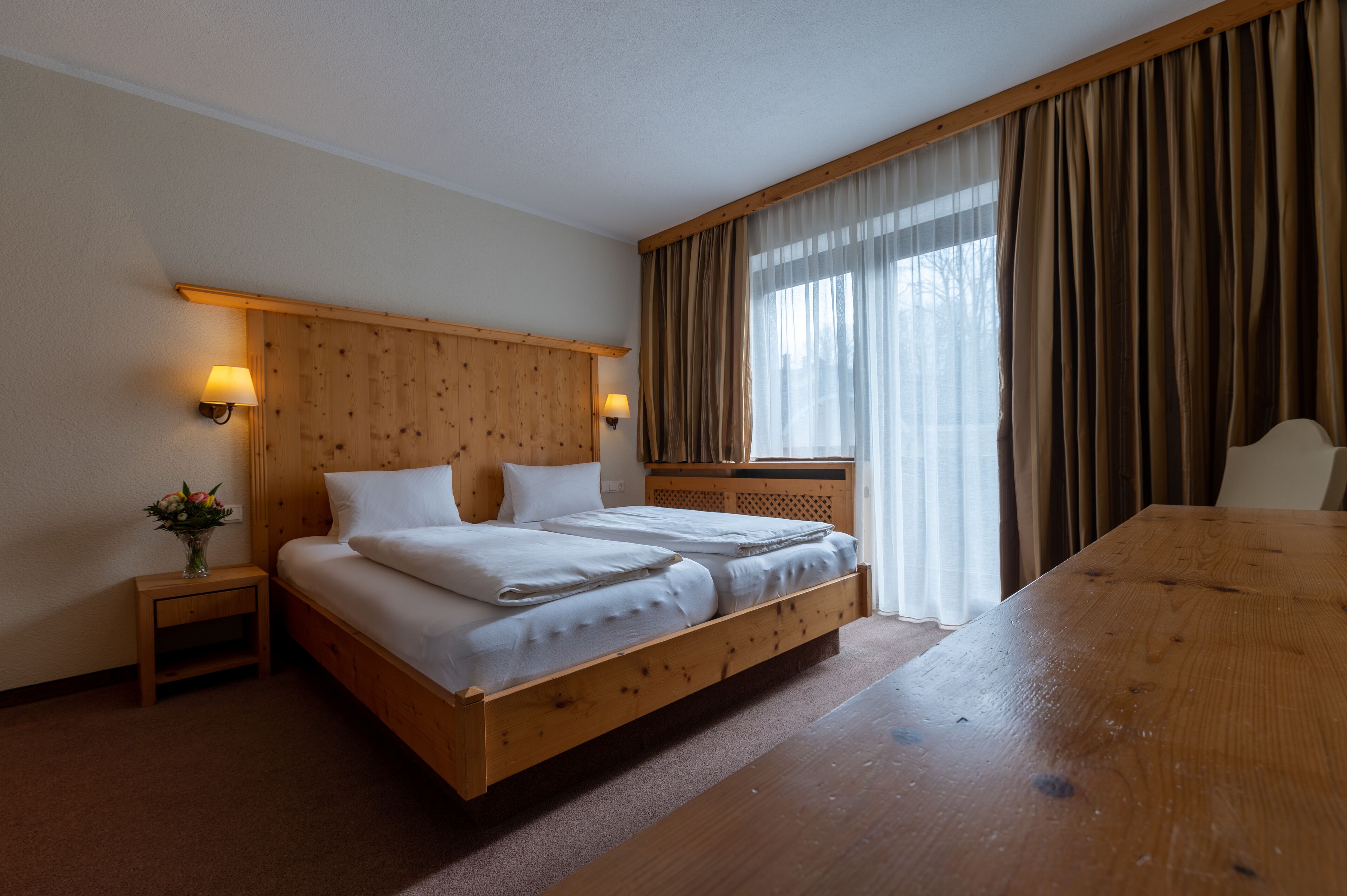 Hotellihuoneessa on puinen sänky valkoisilla lakanoiden kanssa, yöpöytä lampulla ja puinen pöytä.
