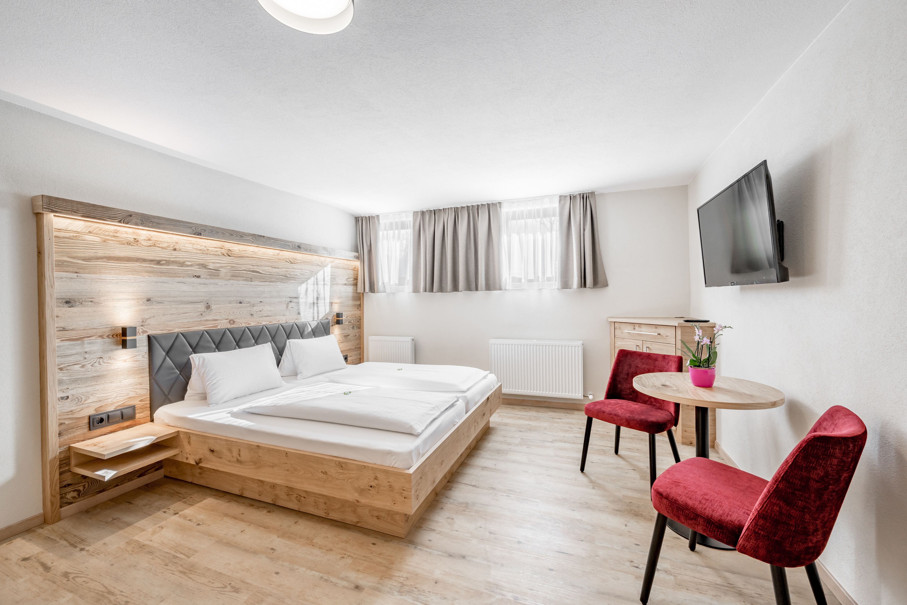 Une chambre moderne avec une tête de lit en bois, un lit, un bureau, deux chaises et un téléviseur fixé au mur.