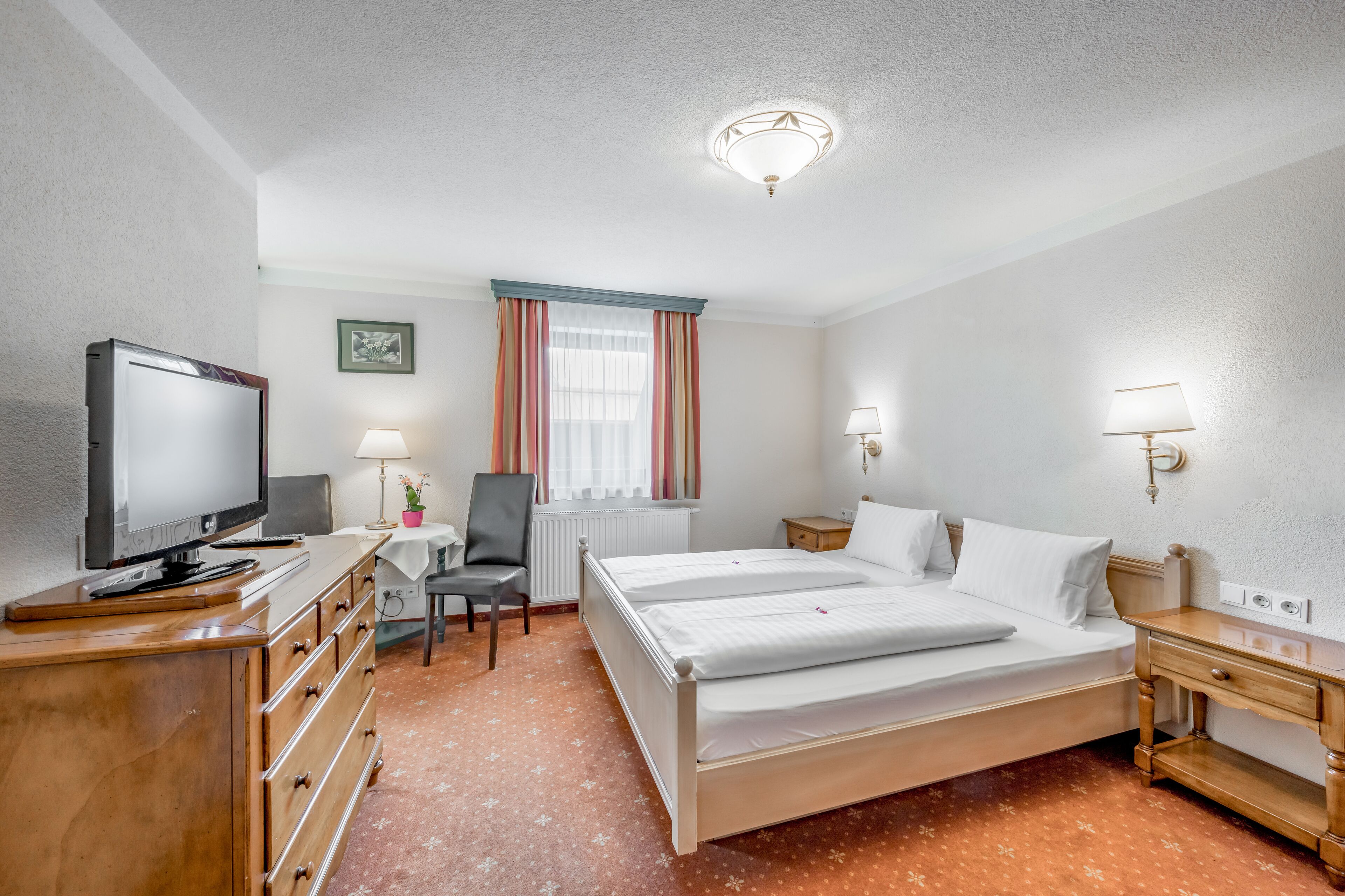 Habitación de hotel con una cama, un escritorio con televisor, una silla, una lámpara y una ventana con cortinas.