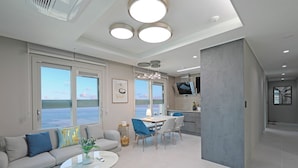 Un salon moderne avec un canapé, une table basse et un coin repas. Une grande fenêtre offre une vue sur la mer.