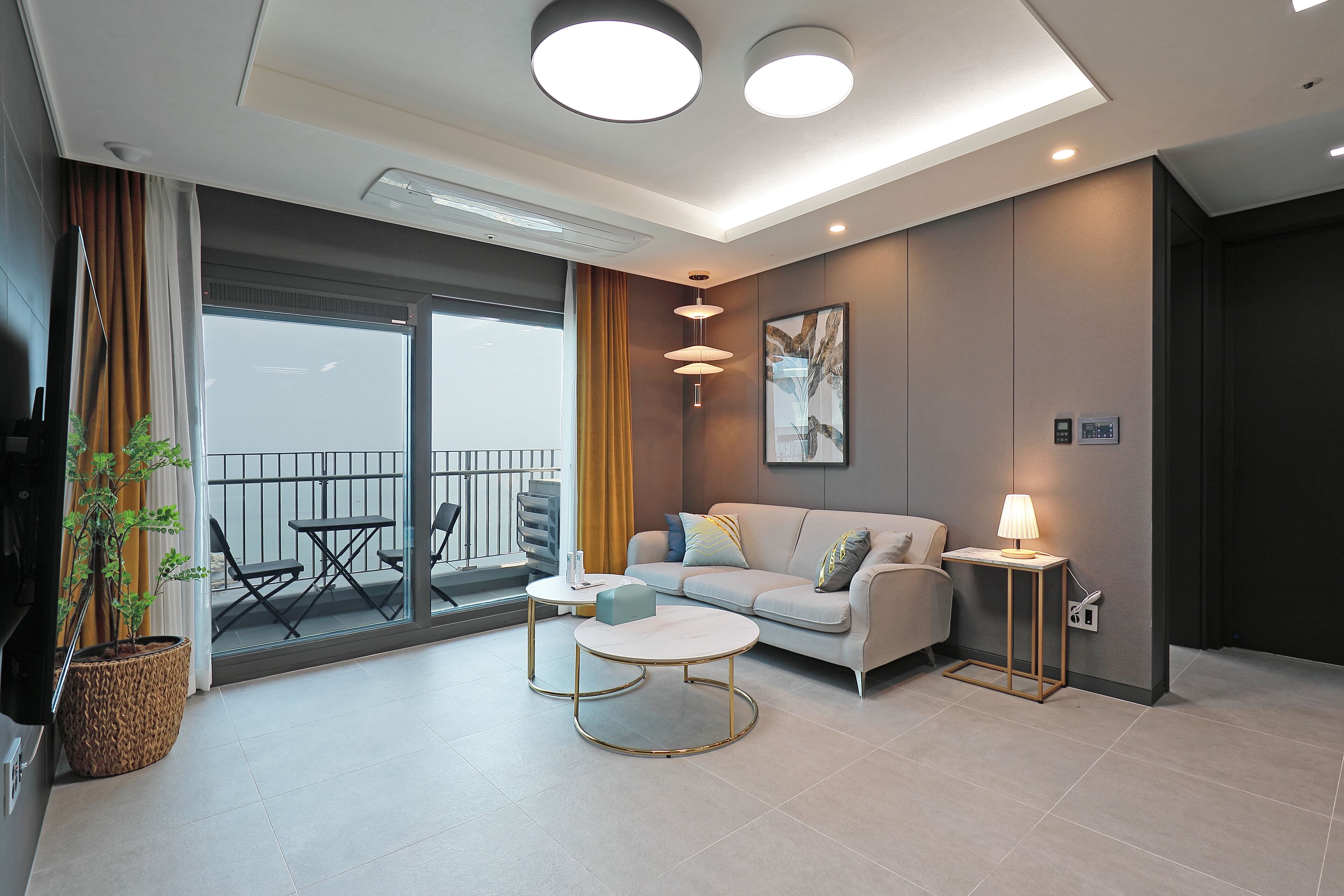 Uma sala de estar moderna com um sofá, mesa de centro e uma luminária. Há uma varanda com uma mesa, cadeiras e um vaso com planta.