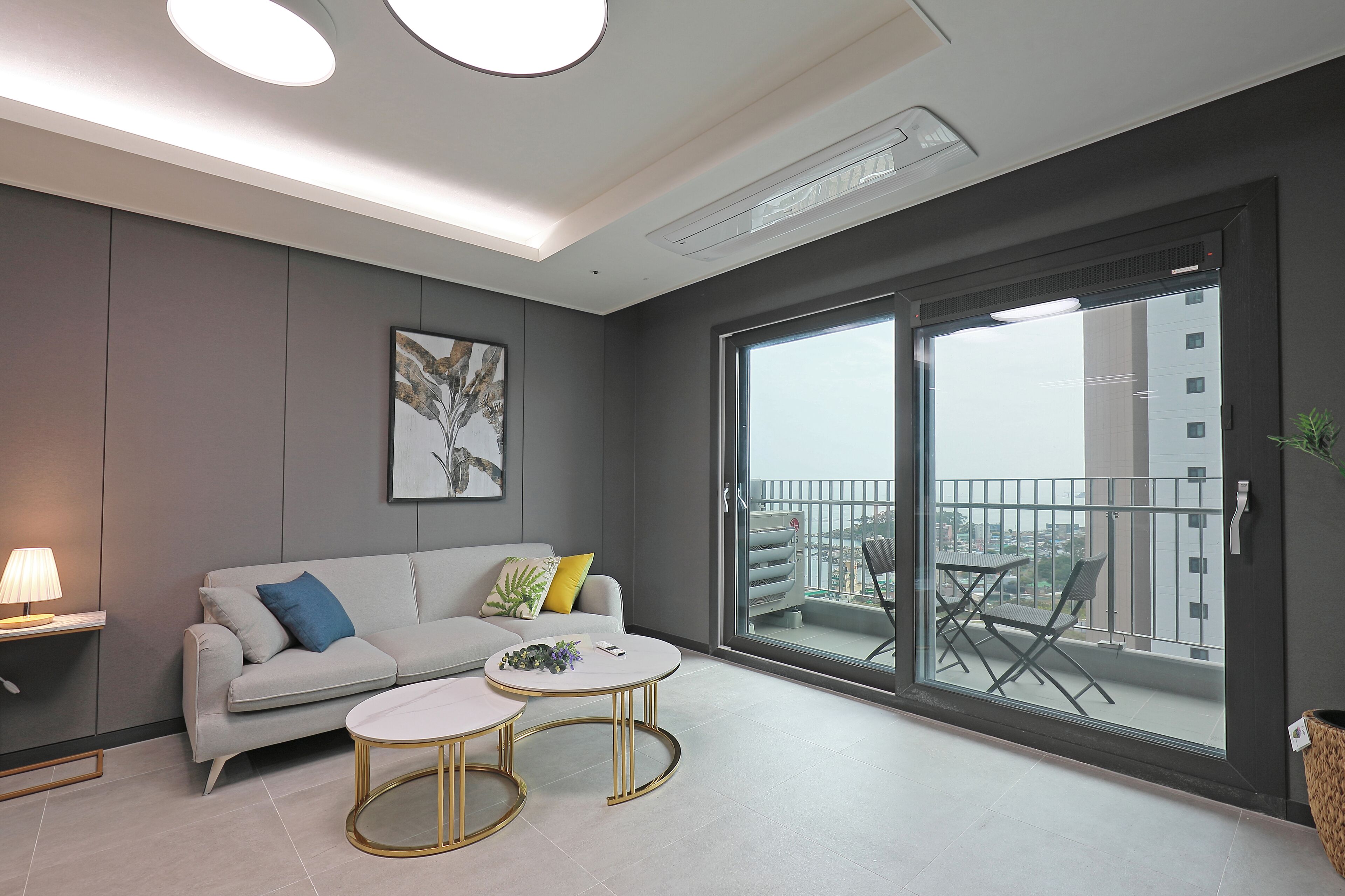 A modern living room with a grey sofa, two white round tables, a floor lamp, and a large sliding glass door leading to a balcony with outdoor furniture.