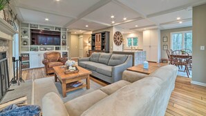 A spacious living room with a grey sofa, beige armchairs, a wooden coffee table, and a fireplace. There is a television, a clock, and a dining area visible in the background. - 2 Mi to Tunnel of Trees: Home w/ Furnished Deck (Harbor Springs)