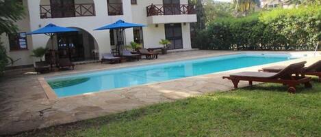 A pool area with lounge chairs and umbrellas, a building with balconies, and a well-maintained lawn.