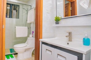 A bathroom with a white toilet, a glass shower enclosure, a sink with a mirror above, and a small potted plant on the counter. - Makao Suite (Nairobi)
