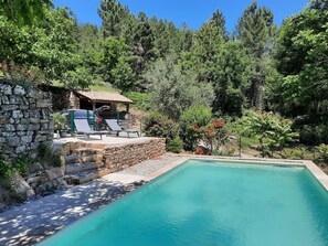 A swimming pool with a stone wall and lounge chairs, surrounded by trees and a small building.