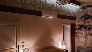Ein Schlafzimmer mit einem Bett, Nachttischlampen, einem Deckenventilator, einer Klimaanlage und einer Wand mit einem Baumgemälde.