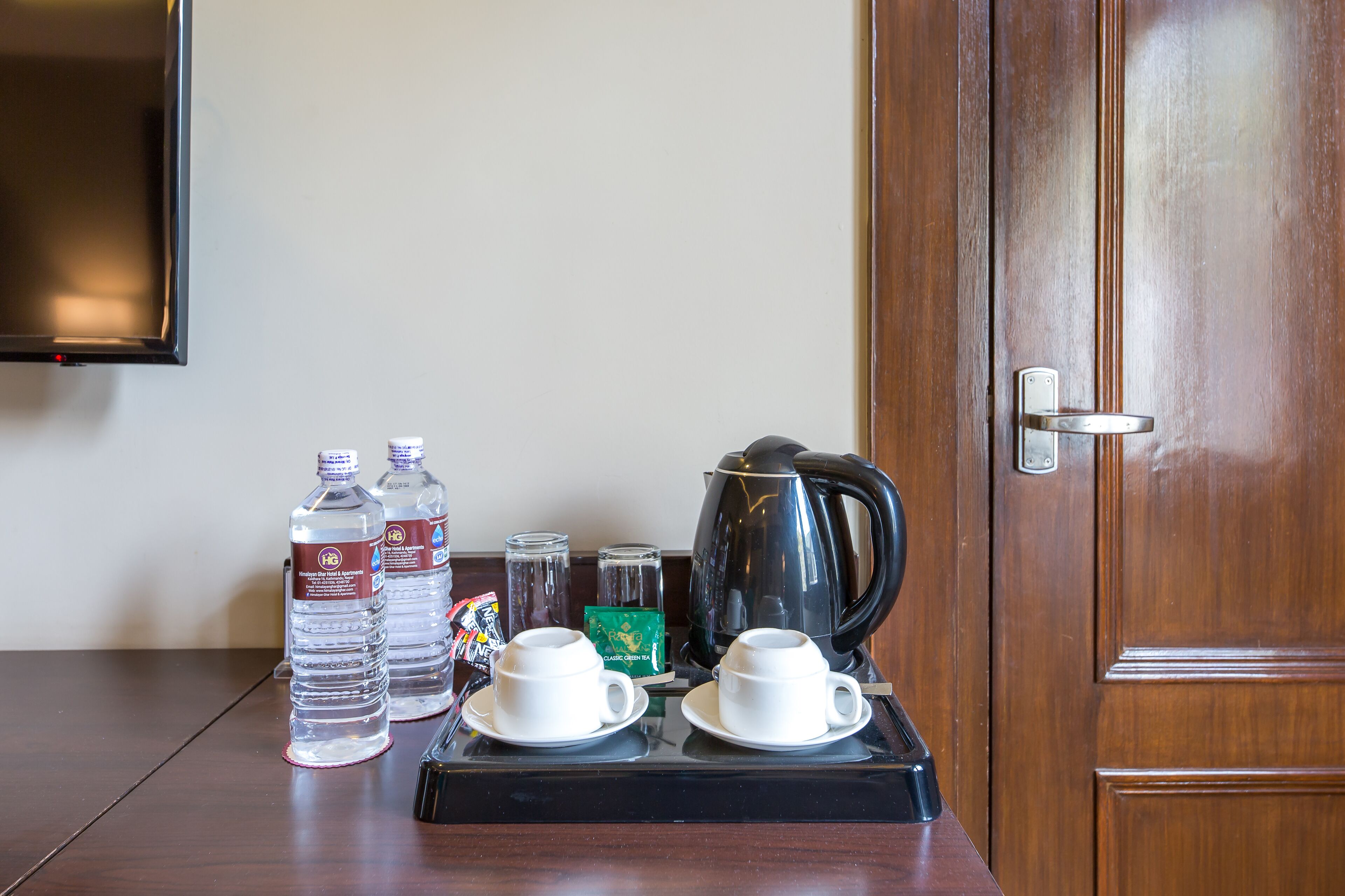 Habitación de hotel con puerta de madera, un televisor de pantalla plana montado en la pared y una mesa con botellas de agua, una cafetera y dos tazas blancas con platillos.