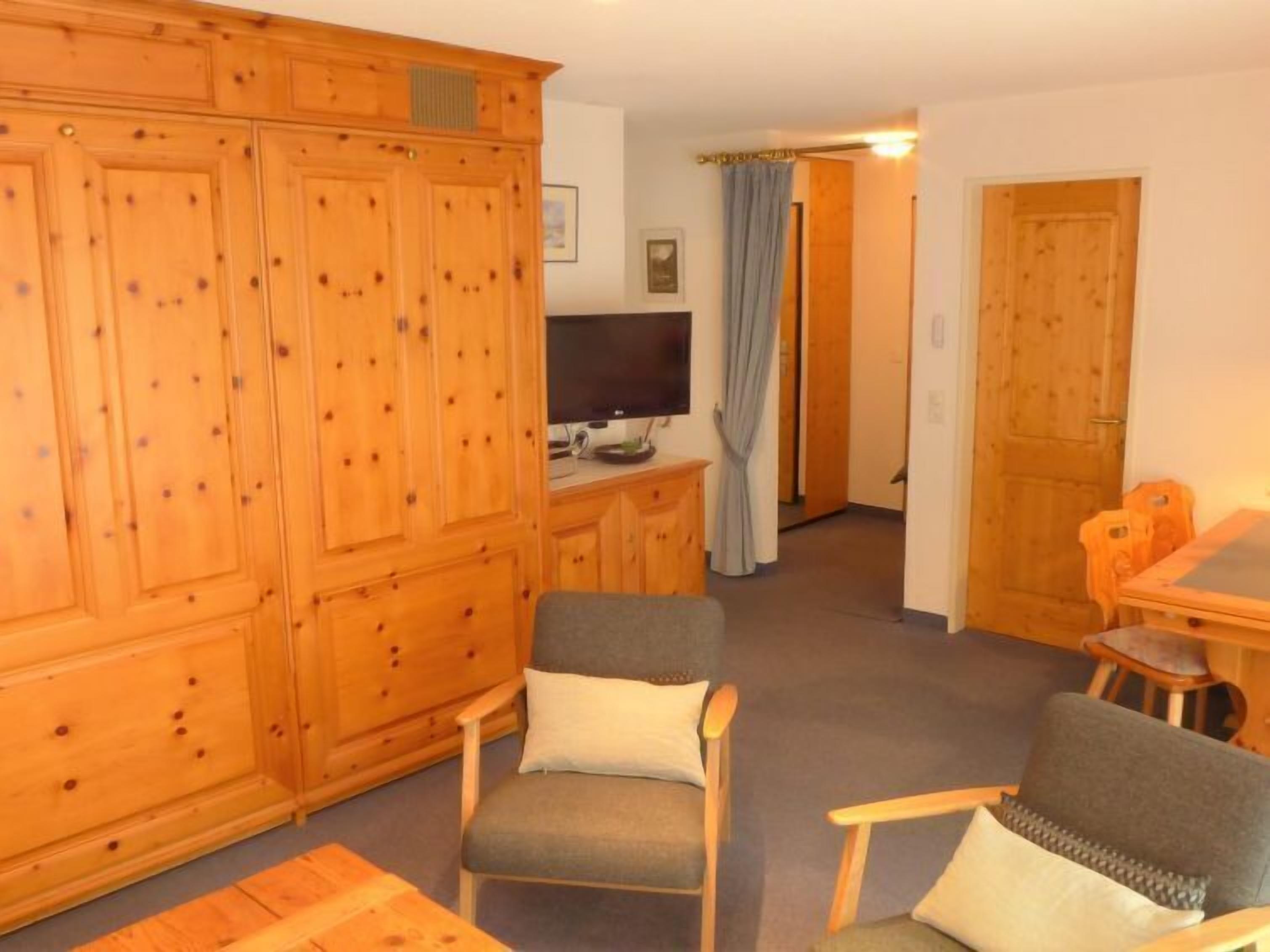 A room with a wooden wardrobe, a television, a grey sofa, and a wooden dining table.