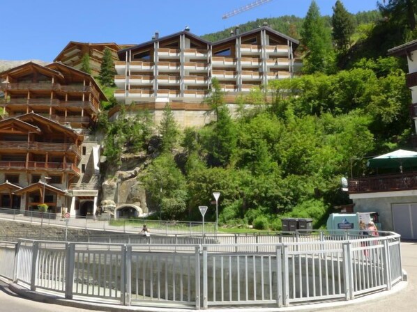 A multi-story wooden building with balconies, set against a backdrop of greenery and a rocky hillside.