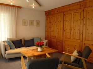 Ein gemütliches Wohnzimmer mit einer Couch, einem Holztisch und einem Sessel. An den Wänden hängen gerahmte Bilder und es gibt einen großen Holzschrank.